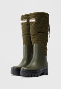 Een paar olijfgroene, gequiltte laarzen tot de knie met zwarte chunky zolen en Barbour-logo patches nabij de bovenkant.