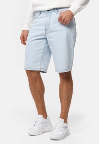 Shorts en denim bleu clair avec une coupe droite, cinq poches et des coutures visibles. Associés à des chaussures de sport blanches.
