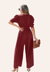 Mono burdeos con mangas abullonadas, cintura fruncida y diseño de pierna ancha. Textura suave, combinado con un bolso de mano marrón y tacones.