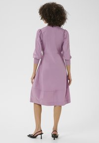 Lila klänning med puffärmar, midjeband och utsvängd kjol. Slät tyg med en subtil glans, som slutar precis ovanför anklarna. Svarta slingback-klackar.