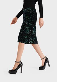 Gonna a matita nera con paillettes, caratterizzata da accenti verdi e blu, abbinata a eleganti scarpe con tacco alto in vernice nera lucida con cinturini alla caviglia.