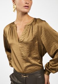 Bruine satijnen blouse met een V-hals en plooien op de schouders, gecombineerd met hoge, donkere broeken met een gouden accentriem.
