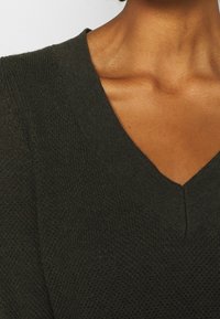 Clavicule et cou d'une personne portant un pull vert foncé en tricot à col en V avec un tissu texturé.
