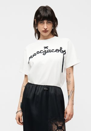 Femeie cu păr șaten până la umeri, purtând un tricou alb cu text negru și o fustă neagră cu un panou din dantelă.