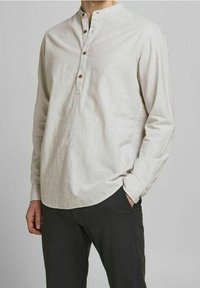 Muž oblečený v béžové dlouhé haleně s třemi knoflíky a černých kalhotách, stojící s jednou rukou v kapse.