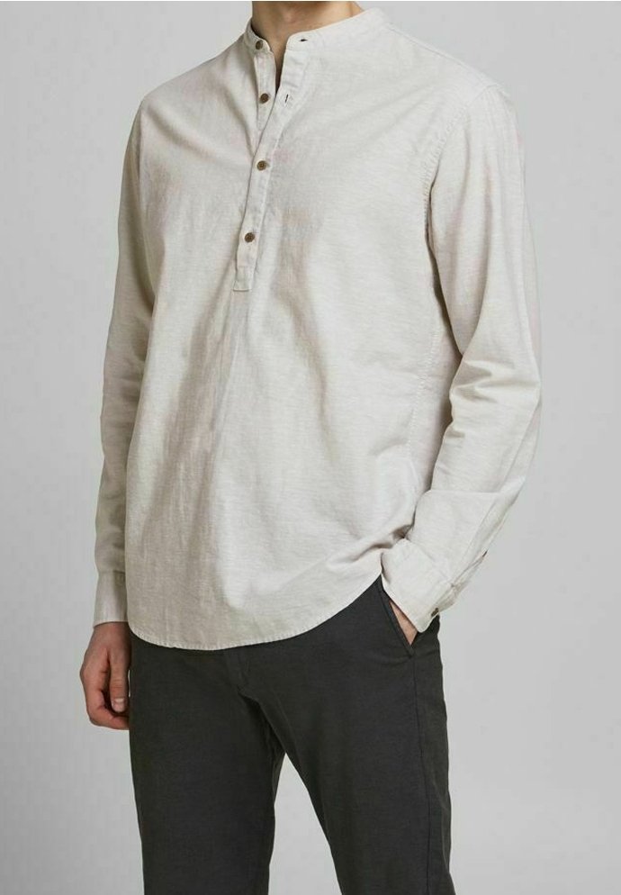 Muž oblečený v béžové dlouhé haleně s třemi knoflíky a černých kalhotách, stojící s jednou rukou v kapse.