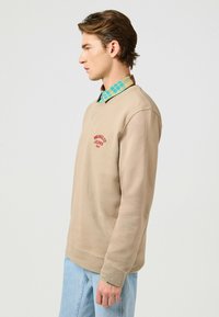 Felpa beige con logo rosso, collo a giro, e polsini a costine, indossata sopra una camicia a quadri verde e blu. Jeans azzurri.