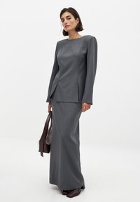 Siva dolg oblek z oblikovanimi rameni, dolgimi rokavi in stranskimi razporki; gladka tkanina in minimalni šivi; kombinirana z rjavim nahrbtnikom.