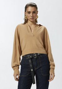 Maglione beige con scollo a V e maniche lunghe, abbinato a jeans in denim scuro a vita alta e una cintura nera con accenti metallici.