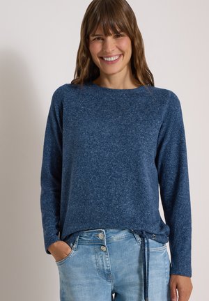 Femme souriante aux cheveux bruns portant un pull bleu à manches longues et un jean bleu clair, les mains dans les poches, sur fond uni.