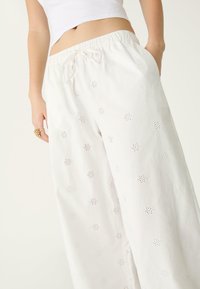 Weiße Baumwollhose mit lockerer Passform, ausgestattet mit Ösenmustern und Taschen. Elastischer Bund mit Kordelzug zur Anpassung.
