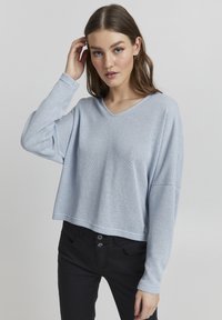 Maglione corto azzurro chiaro, texturizzato, con scollo a V e maniche lunghe, abbinato a pantaloni neri con dettagli di bottoni.