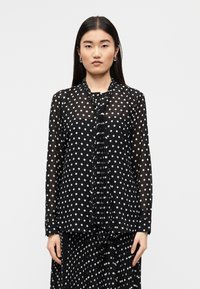 Μαύρο διάφανο μπλουζάκι με λευκά πουά, μακριά μανίκια και πιετέ μπροστινή δέστρα. Το ύφασμα φαίνεται ελαφρύ με λεία υφή.