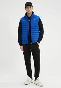 Niebieska kamizelka puchowa z zamkiem z przodu, poziomym pikowaniem i akcentami logo; nałożona na czarny długi rękaw i czarne spodnie, z białymi sneakersami.