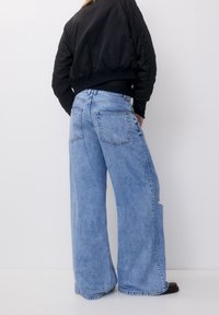 Jean large bleu clair avec une finition délavée, doté de poches avant et arrière ainsi qu'un détail usé. Associé à une veste bomber noire.