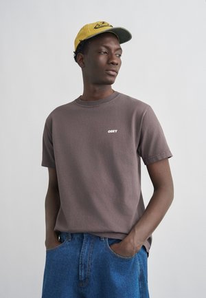 Fiatal férfi sárga sapkában, szürke OBEY pólóban és kék farmerben, kezét a zsebében tartva, oldalra néz egy egyszerű háttér előtt.