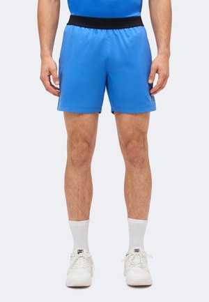 Homme portant un short de sport bleu vif avec une ceinture noire, des chaussettes blanches hautes et des baskets blanches, debout devant un fond uni.
