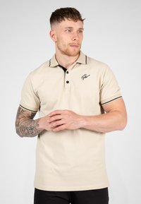 Beige poloshirt met zwarte kraagaccenten en zoom, voorzien van een drieknoopsluiting en een klein geborduurd logo op de borst.