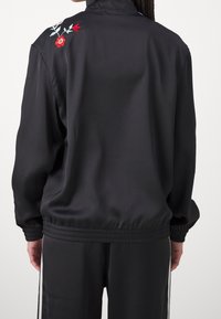 Svart idrottsjacka med hög krage, prydd med röd och grön blommönsterbroderi på baksidan, tillverkad av mjukt, lättviktigt tyg.