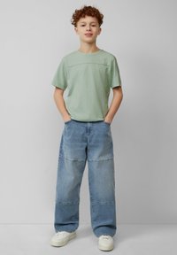 Lichtgroene T-shirt met korte mouwen en naaddetails, gecombineerd met losvallende blauwe denim jeans met een patchworkontwerp. Witte sneakers maken de look af.