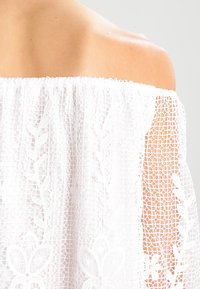 Top bianco scollato sulle spalle realizzato in pizzo con motivi floreali e maniche corte semi-trasparenti. Dettagli intricati lungo l'orlo.