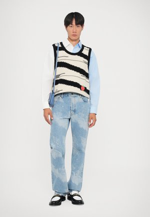 Chaleco de punto con patrón de zigzag en negro y crema, camisa azul claro, jeans azul desgastado con patrón de nubes y zapatos negros con acentos blancos.