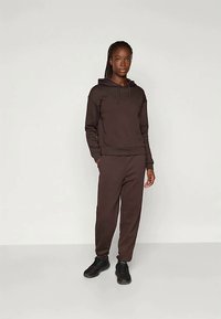 Rjava kapuca v kombinaciji s usklajenimi trenirkami, izdelanimi iz mehkih materialov. Ima nastavljiv kapuco in rebraste manšete. Črna obutev dopolnjuje videz.