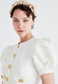 Λευκή μπλούζα με φουσκωτά μανίκια, η οποία διαθέτει περίτεχνους χρυσούς και μαργαριταρένιους κουμπιά, υφή στο ύφασμα και στρογγυλή λαιμόκοψη. Συμπληρώνεται με μπεζ κορδέλα για το κεφάλι.