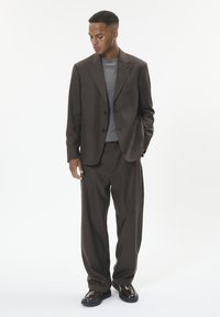 Dark brown suit with a tailored fit, paired with a gray t-shirt. Features flat front pants and shiny black shoes. Plain background.