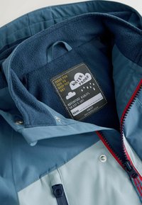 Inderside af vandtæt jakke med blødt blå fleecefor, en mærkat med teksten "VANDTÆT" og kontrasterende rød lynlås samt detaljer.