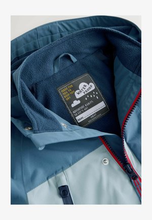 Vattentät jackas insida med mjukt blått fleecefoder, en etikett med texten "VATTENTÄT" och kontrasterande röd dragkedja och detaljer.