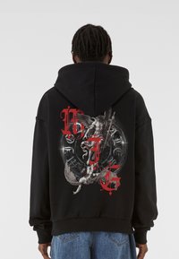 Hanorac negru cu un grafic mare și detaliat pe spate, cu text roșu și designuri intricate ale unei figuri cu aripi pe un fundal circular.