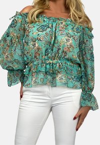 Lätt, off-the-shoulder blus i akvamarinfärg med paisleymönster. Har volangärmar och resårmidja. Matchas med vita byxor.