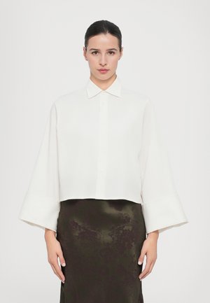 Cropped white blouse with a collar and wide sleeves, made from a smooth fabric. Paired with a dark green textured floral skirt.