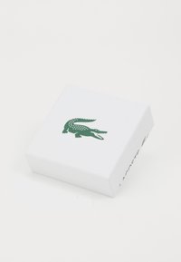 Hvid rektangulær kasse med en tekstureret overflade, der har et grønt krokodille-logo på toppen og "LACOSTE" trykt på den ene side.
