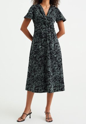 Frau trägt ein schwarzes Midikleid mit grünen Blattmustern, kurzen Ärmeln, V-Ausschnitt, verdrehtem Vorderteil und schwarzen, geschnürten Sandaletten mit Absatz.