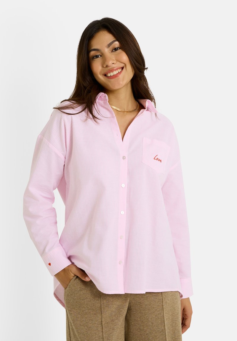 Chemise boutonnée rose clair avec des rayures blanches discrètes, dotée d'une petite poche avec le mot "Love" brodé en rouge, et des manches à coupe décontractée.