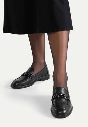 Schwarze Lederloafer mit einer Kettenakzentuierung an der Vorderseite. Flache Sohlen mit einer abgerundeten Zehenform und niedrigem Absatz. Getragen mit transparenten schwarzen Strumpfhosen.