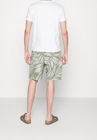 Lettere hvid t-shirt, afslappet pasform. Olivenfarvede shorts med stort hvidt bladmønster. Brune sandaler. Udsigt fra bagsiden.