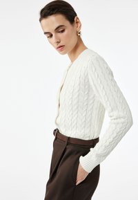 Weißer Strickcardigan mit Rippbund und Rippärmeln, kombiniert mit braunen hoch taillierten Hosen. Mit einer Knopfverschluss-Front.