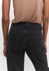 Jeans de mezclilla negra con tiro alto, bolsillos traseros visibles y detalles en costura verde; combinados con una camiseta sin mangas marrón.