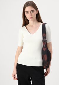 Weißes, geripptes Kurzarmoberteil mit V-Ausschnitt. Das Modell hält außerdem eine dunkelrote und schwarz gemusterte Tasche mit einem Schmalen, metallbeschlagenen Trageriemen.