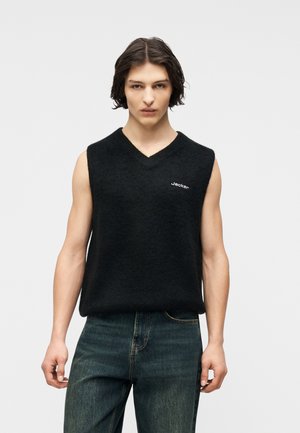 Junger Mann mit mittellangem dunklem Haar trägt einen schwarzen ärmellosen V-Ausschnitt-Pullover mit dem Schriftzug "Jacken" und dunkelblaue Jeans vor grauem Hintergrund.