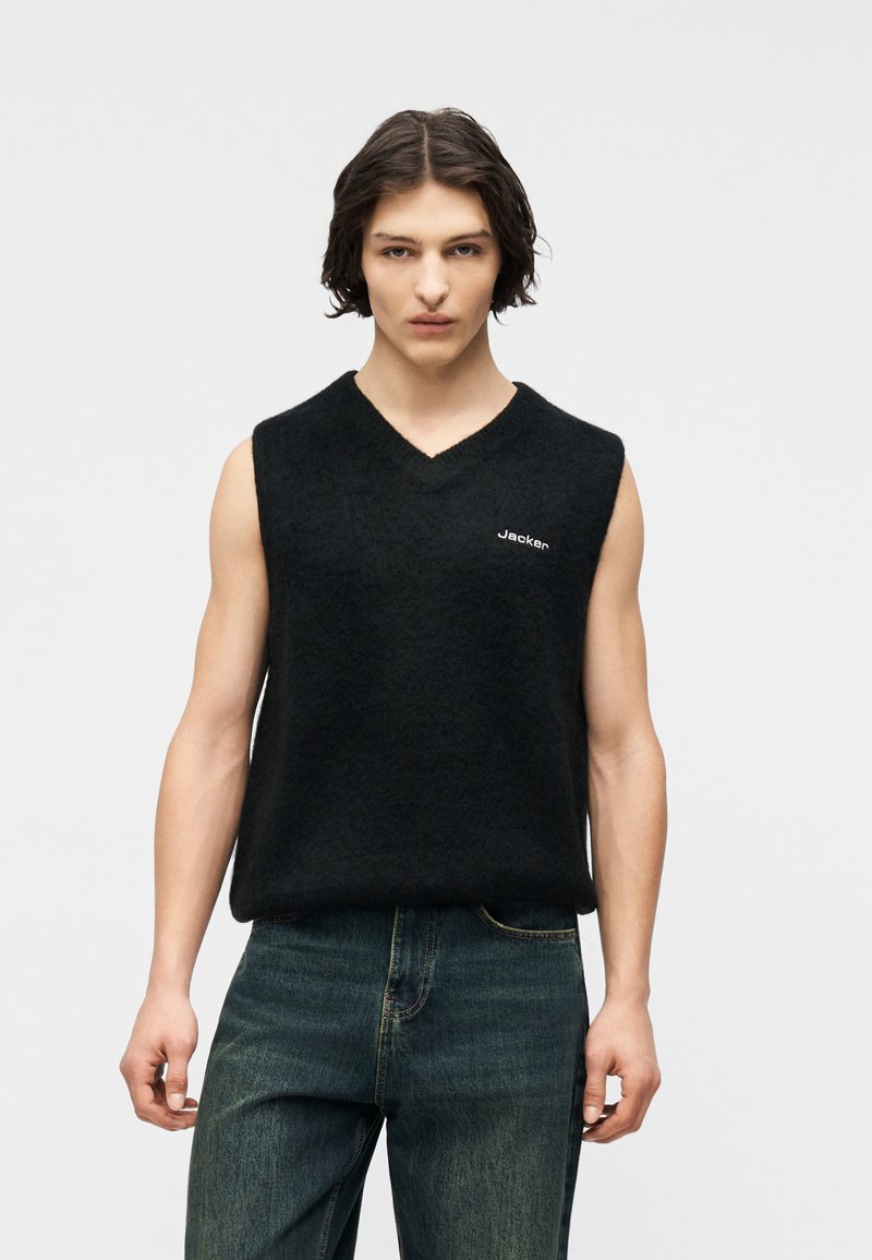 Νεαρός άνδρας με μεσαίου μήκους σκουρόχρωμα μαλλιά φοράει μαύρο αμάνικο πουλόβερ με V-λαιμόκοψη και το κείμενο "Jacken" καθώς και σκούρο μπλε τζιν, σε φόντο γκρι.