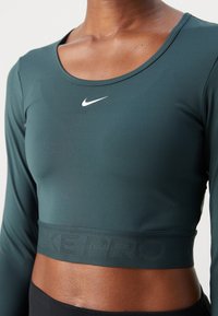 Top scurt cu mânecă lungă de culoare verde închis, fabricat dintr-un material neted, având un logo alb Nike și un elastic texturat de culoare neagră cu textul "NIKE PRO".