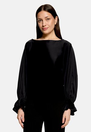 Vrouw met lang donker haar die een zwarte satijnen blouse draagt met lange gepofde mouwen en gerimpelde manchetten, staand tegen een effen lichte achtergrond.