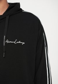 Fekete kapucnis pulóver, fehér "Armani Exchange" felirattal a mellet, illeszkedő márkás zsinórral és fehér csíkokkal az ujjak mentén.