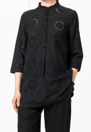 Woman wearing a black long-sleeve blouse with circular eyelet patterns and buttons down the front, paired with black pants.