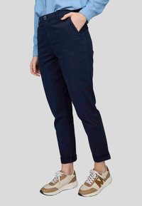 Pantalon bleu marine avec une texture lisse, présentant une fermeture à bouton, des poches latérales et des ourlets repliés, associé à des baskets beige clair et blanches.