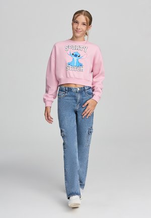 Felpa rosa corta con un personaggio dei cartoni animati blu e la scritta "SPORTY STITCH". Abbinata a jeans baggy azzurri e sneakers bianche.